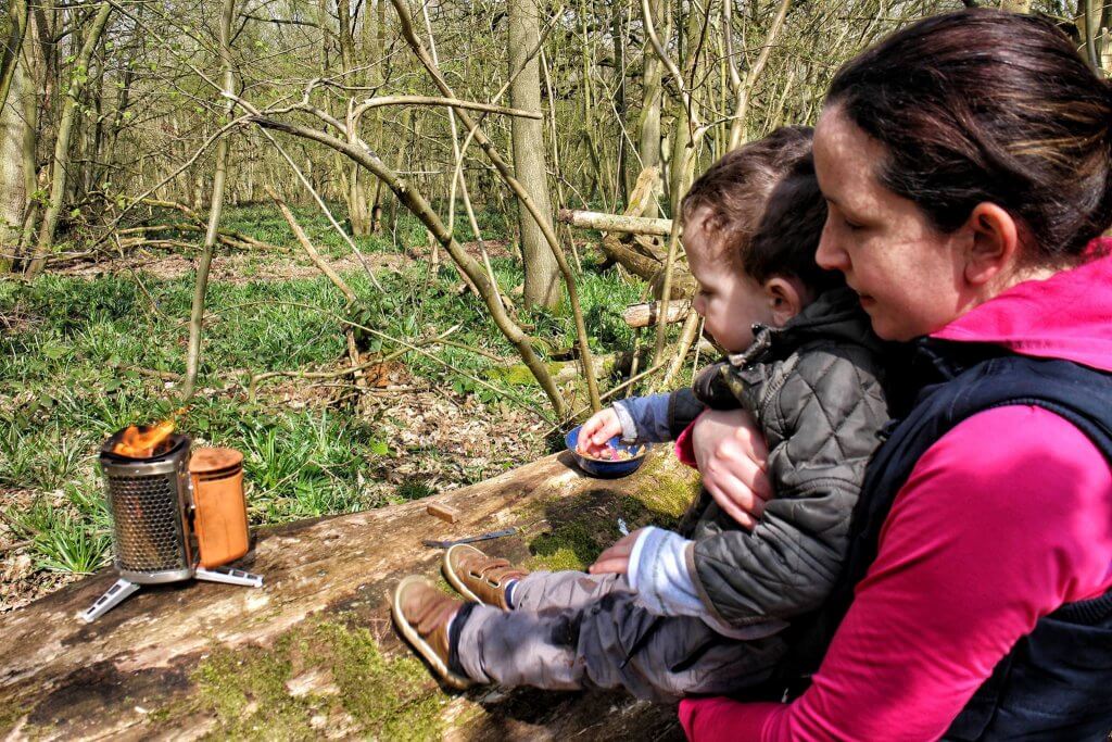 A woman and toddler look at a camping stove. They are in a forest